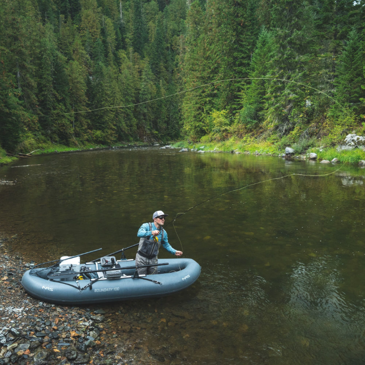 Professional fly fishing guide demonstrating proper casting stance and grip with a fly rod while standing in shallow river water, showing arm position and body alignment with clear water backdrop